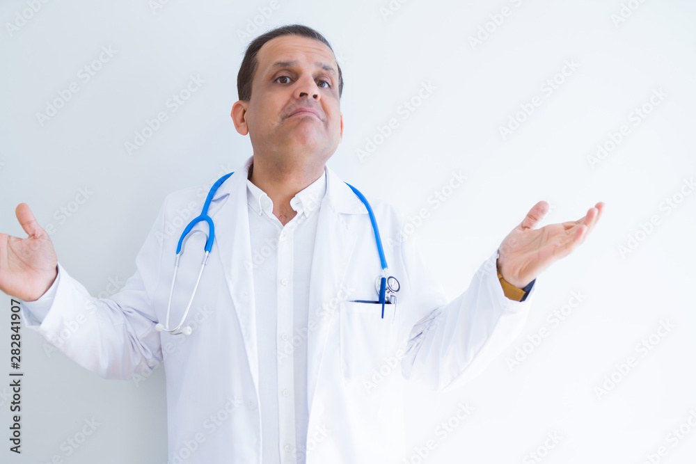 Middle age doctor man wearing stethoscope and medical coat over white background clueless and confused expression with arms and hands raised. Doubt concept. Middle age doctor man wearing stethoscope and medical coat over white background clueless and confused expression with arms and hands raised. Doubt concept.