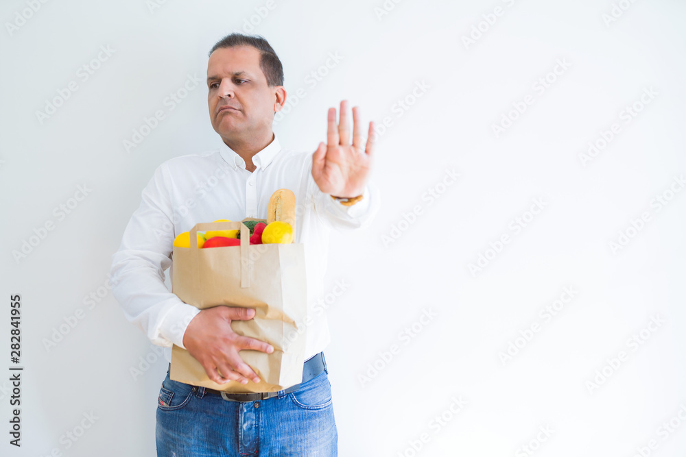Middle age man holding groceries shopping bag over white background with open hand doing stop sign with serious and confident expression, defense gesture Middle age man holding groceries shopping bag over white background with open hand doing stop sign with serious and confident expression, defense gesture