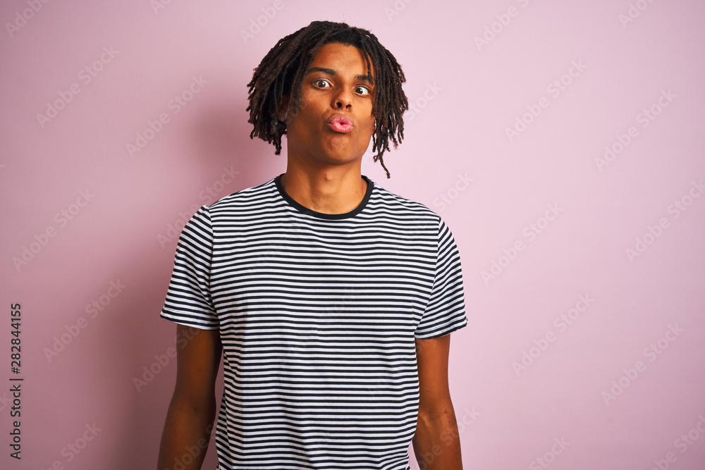 Afro man with dreadlocks wearing navy striped t-shirt standing over isolated pink background making fish face with lips, crazy and comical gesture. Funny expression. Afro man with dreadlocks wearing navy striped t-shirt standing over isolated pink background making fish face with lips, crazy and comical gesture. Funny expression.
