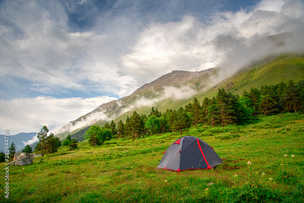 tourist tent stands on a green meadow against the backdrop of mountain slopes and peaks with clouds and blue sky
