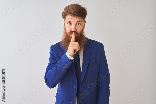 Young redhead irish businessman wearing suit standing over isolated white background asking to be quiet with finger on lips. Silence and secret concept.