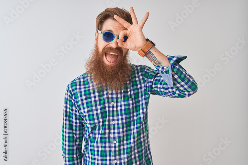 Young redhead irish man wearing casual shirt and sunglasses over isolated white background doing ok gesture with hand smiling, eye looking through fingers with happy face.