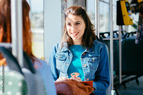 Two cheerful pretty young women are sitting in front of each other in a bus or tram and looking at the books, reading, talking smiling while waiting for a bus to take them to their destination.