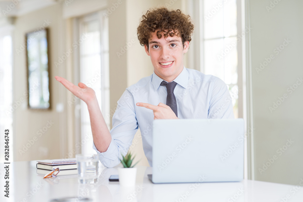 Young business man working with computer laptop at the office amazed and smiling to the camera while presenting with hand and pointing with finger.