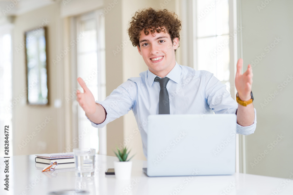 Young business man working with computer laptop at the office looking at the camera smiling with open arms for hug. Cheerful expression embracing happiness.