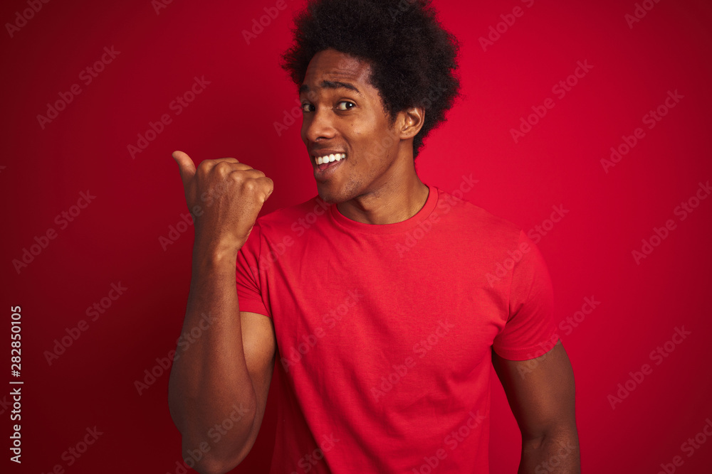 Young american man with afro hair wearing t-shirt standing over isolated red background smiling with happy face looking and pointing to the side with thumb up. Young american man with afro hair wearing t-shirt standing over isolated red background smiling with happy face looking and pointing to the side with thumb up.