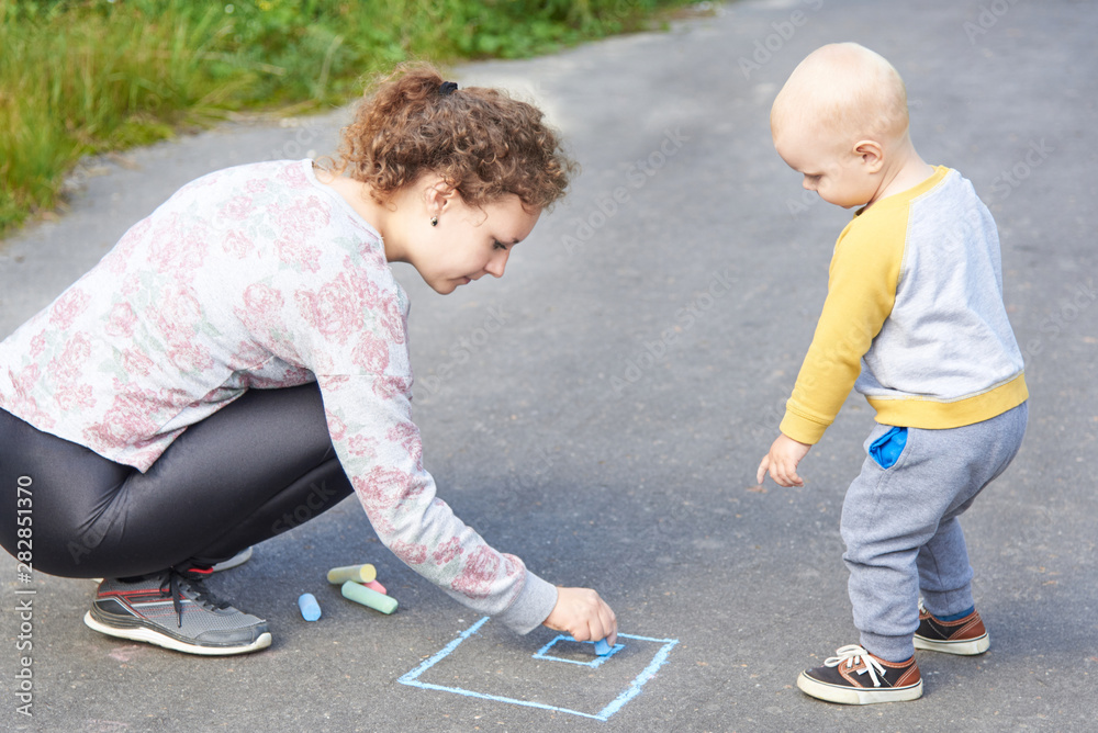 Fototapeta premium Mother with her little son draws a colorful chalk on asphalt on a summer day. Mom and child activity.