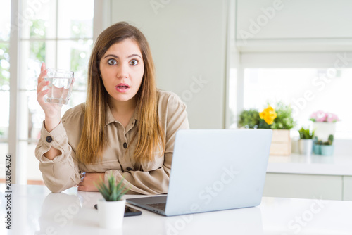 Beautiful young woman working with computer takes a break to drink glass of water scared in shock with a surprise face, afraid and excited with fear expression