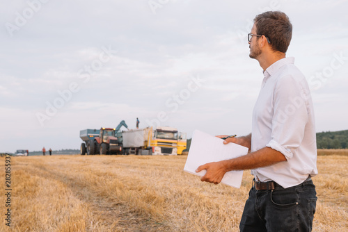 Young agronomist man standing on wheat field checking quality while combine harvester working.