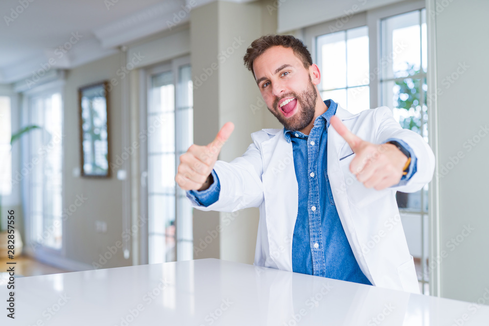 Handsome doctor man wearing medical coat at the clinic approving doing positive gesture with hand, thumbs up smiling and happy for success. Looking at the camera, winner gesture. Handsome doctor man wearing medical coat at the clinic approving doing positive gesture with hand, thumbs up smiling and happy for success. Looking at the camera, winner gesture.