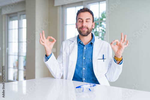 Handsome scientist man wearing white robe and safety glasses relax and smiling with eyes closed doing meditation gesture with fingers. Yoga concept.