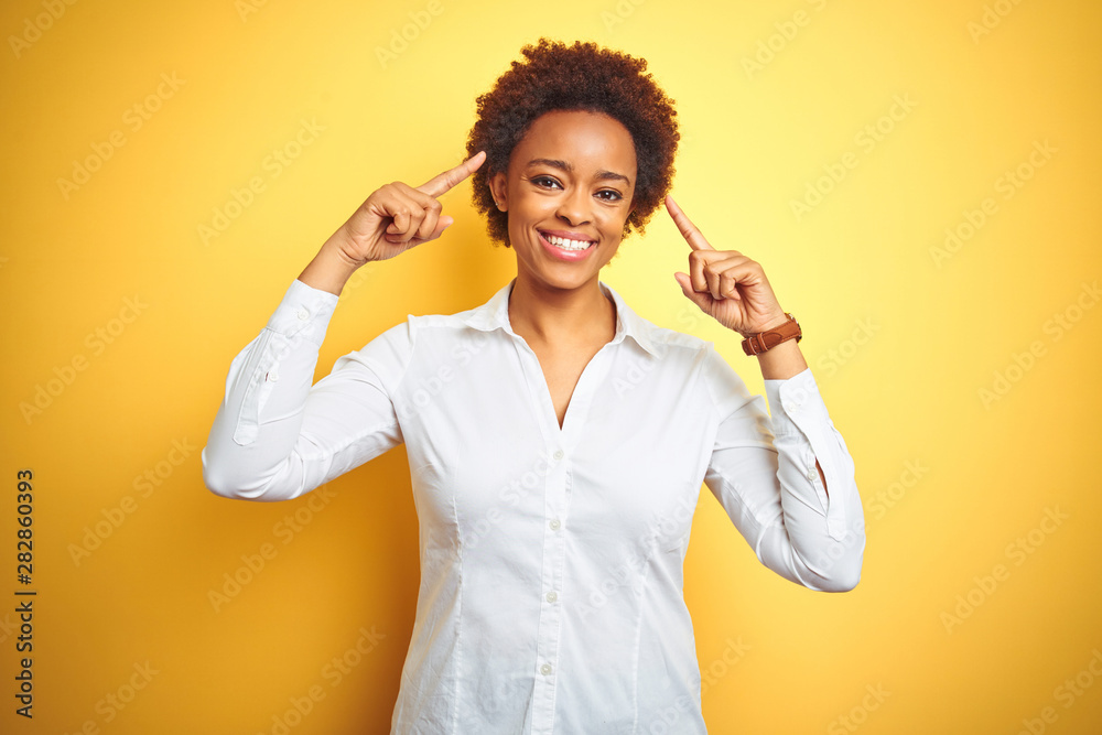 African american business woman over isolated yellow background smiling pointing to head with both hands finger, great idea or thought, good memory African american business woman over isolated yellow background smiling pointing to head with both hands finger, great idea or thought, good memory