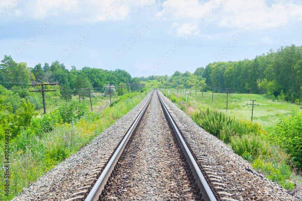 A railway through the summer green fields.  Beautiful green railway tree landscape. White sky. Beautiful landscape. Railway transport.