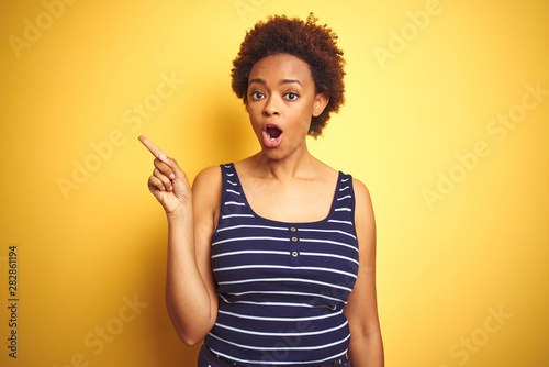 Beauitul african american woman wearing summer t-shirt over isolated yellow background Surprised pointing with finger to the side, open mouth amazed expression.