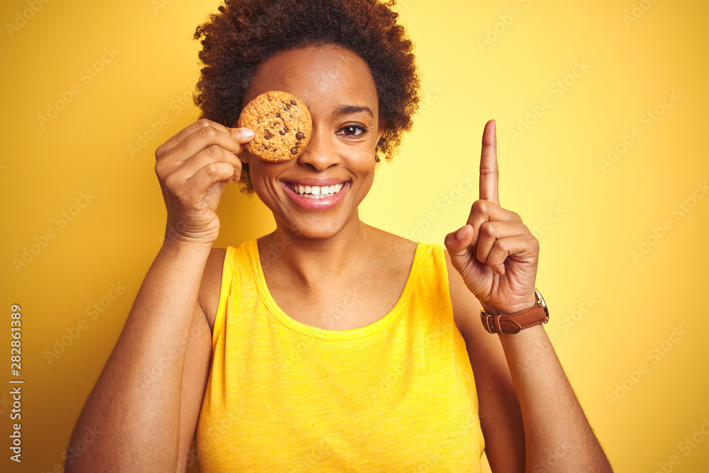 African american woman holding chocolate chips cookie over yellow background surprised with an idea or question pointing finger with happy face, number one African american woman holding chocolate chips cookie over yellow background surprised with an idea or question pointing finger with happy face, number one