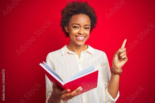 African american woman reading a book over red isolated background very happy pointing with hand and finger to the side