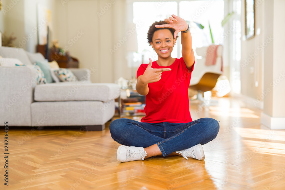 Young beautiful african american woman sitting on the floor at home smiling making frame with hands and fingers with happy face. Creativity and photography concept.
