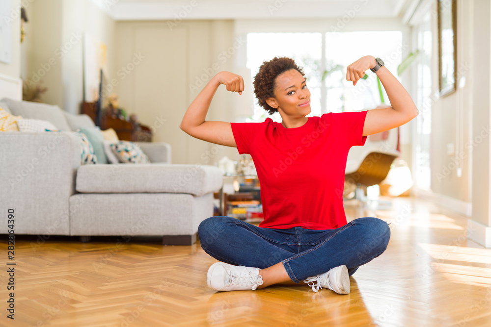 Young beautiful african american woman sitting on the floor at home showing arms muscles smiling proud. Fitness concept.