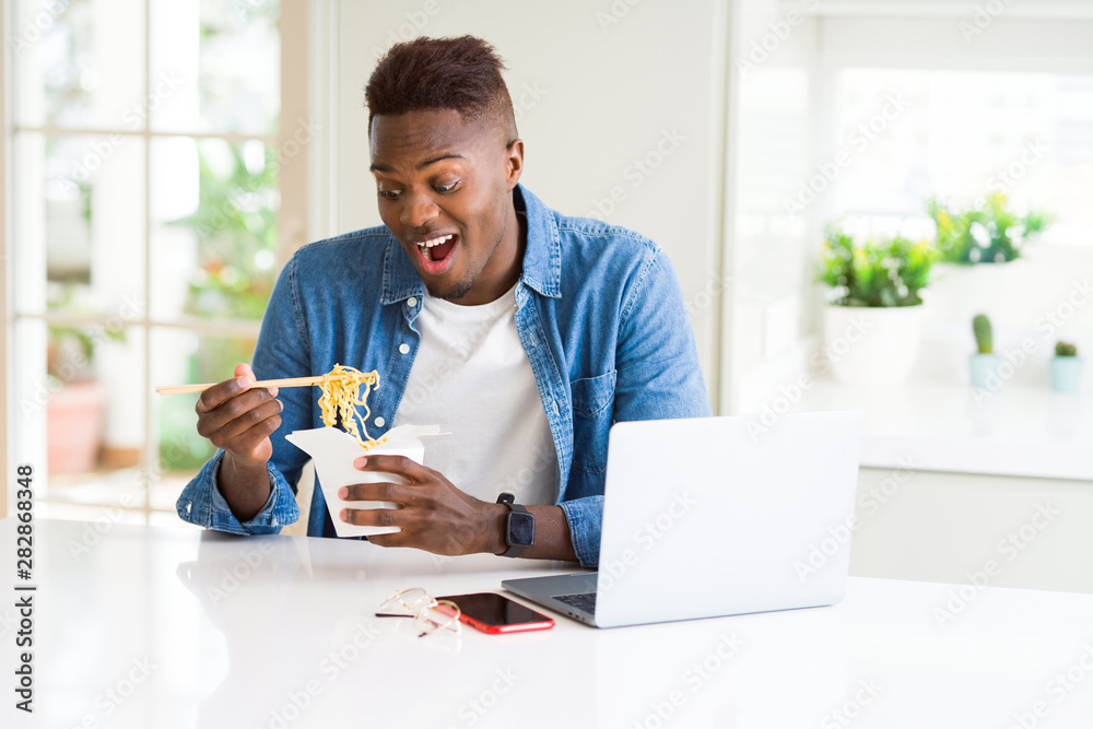 Handsome young african business man eating delivery asian food and working using computer, enjoying noodles smiling Handsome young african business man eating delivery asian food and working using computer, enjoying noodles smiling