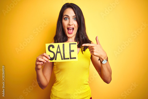 Young beautiful woman holding sale poster over yellow isolated background very happy pointing with hand and finger