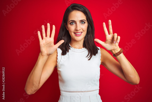 Young beautiful woman wearing white dress standing over red isolated background showing and pointing up with fingers number eight while smiling confident and happy.