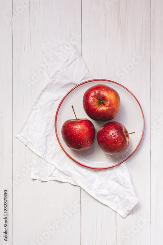 Three ripe red apples on a white plate. Top view on wooden table with a napkin.