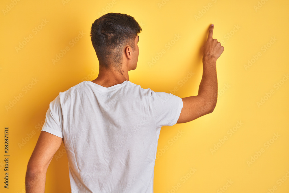 Young indian man wearing white t-shirt standing over isolated yellow background Posing backwards pointing ahead with finger hand Young indian man wearing white t-shirt standing over isolated yellow background Posing backwards pointing ahead with finger hand