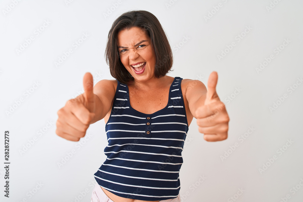 Young beautiful woman wearing blue striped t-shirt standing over isolated white background approving doing positive gesture with hand, thumbs up smiling and happy for success. Winner gesture. Young beautiful woman wearing blue striped t-shirt standing over isolated white background approving doing positive gesture with hand, thumbs up smiling and happy for success. Winner gesture.