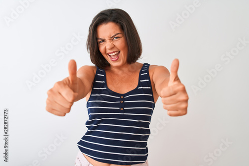 Young beautiful woman wearing blue striped t-shirt standing over isolated white background approving doing positive gesture with hand, thumbs up smiling and happy for success. Winner gesture. © Krakenimages.com