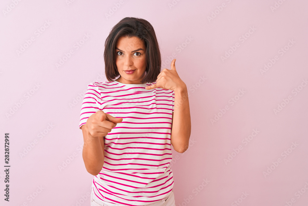Young beautiful woman wearing striped t-shirt standing over isolated pink background smiling doing talking on the telephone gesture and pointing to you. Call me. Young beautiful woman wearing striped t-shirt standing over isolated pink background smiling doing talking on the telephone gesture and pointing to you. Call me.