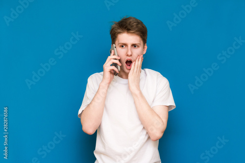 A young man in a white T-shirt holds the phone in his hands with a surprised face and looks down at the side on a blue background. Blue background.