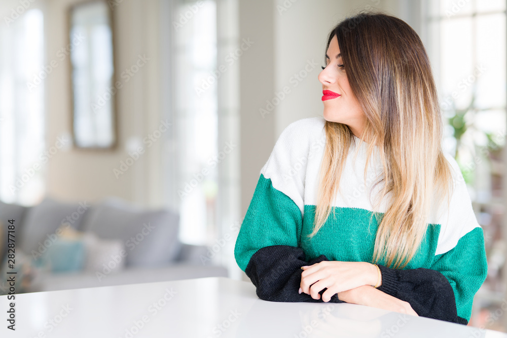 Young beautiful woman wearing winter sweater at home smiling looking to the side with arms crossed convinced and confident