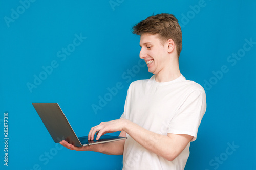 Portrait of a cheerful man, standing on a blue background, holding a laptop in his hands, playing a game. A man with a laptop, wearing a white T-shirt, is isolated on a blue background.