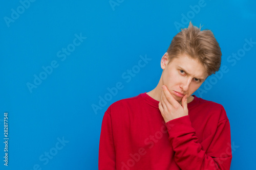 A close-up portrait of a guy standing sad on a blue background and holding his face and looking at the camera. Young sad guy stands on a blue background
