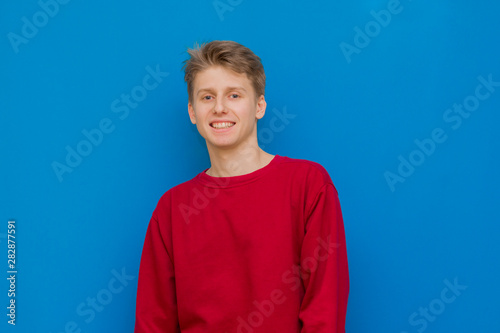 Portrait of a cheerful young guy standing on a blue background and smiling while looking at the camera. Young handsome guy in a red sweater stands on a blue background