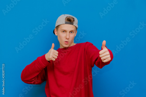 A smiling boy is standing on a blue background, looking at the camera and showing a thumbs up. Positive young man shows a gesture, like, isolated on a blue background,