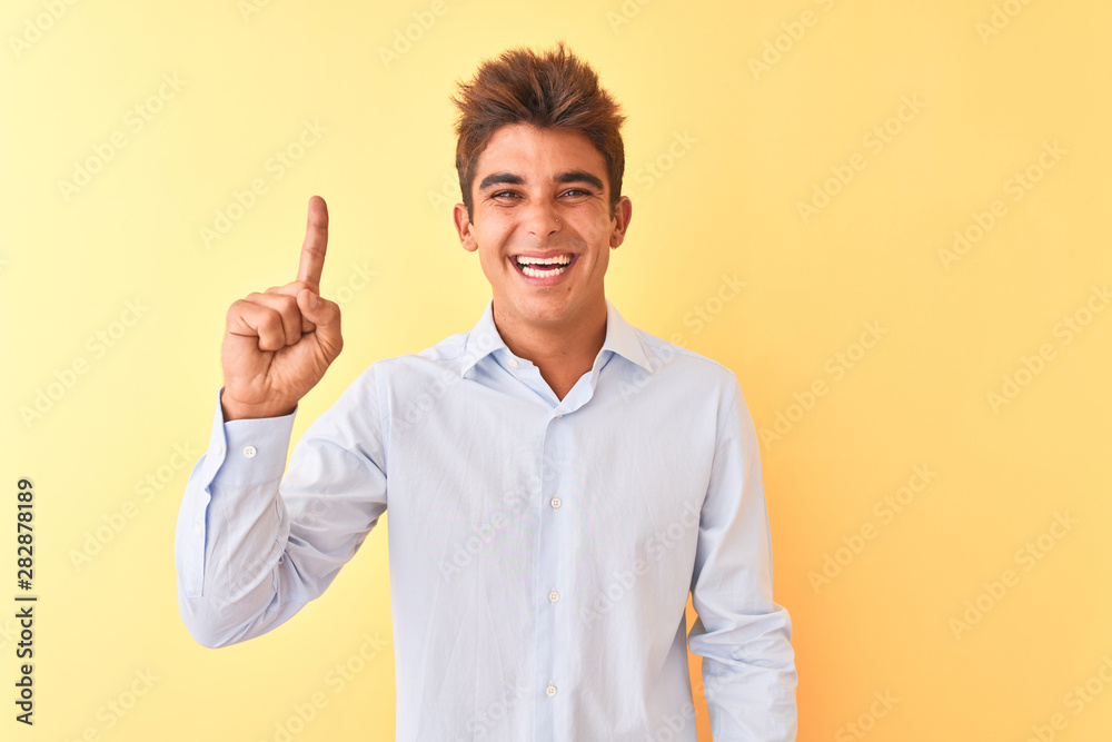 Young handsome businessman wearing elegant shirt over isolated yellow background showing and pointing up with finger number one while smiling confident and happy.