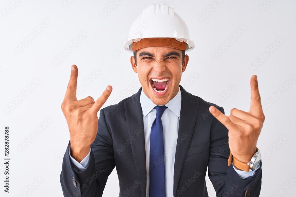 Young handsome architect man wearing suit and helmet over isolated white background shouting with crazy expression doing rock symbol with hands up. Music star. Heavy concept.