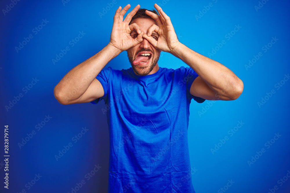 Young handsome man wearing casual t-shirt over blue isolated background doing ok gesture like binoculars sticking tongue out, eyes looking through fingers. Crazy expression. Young handsome man wearing casual t-shirt over blue isolated background doing ok gesture like binoculars sticking tongue out, eyes looking through fingers. Crazy expression.