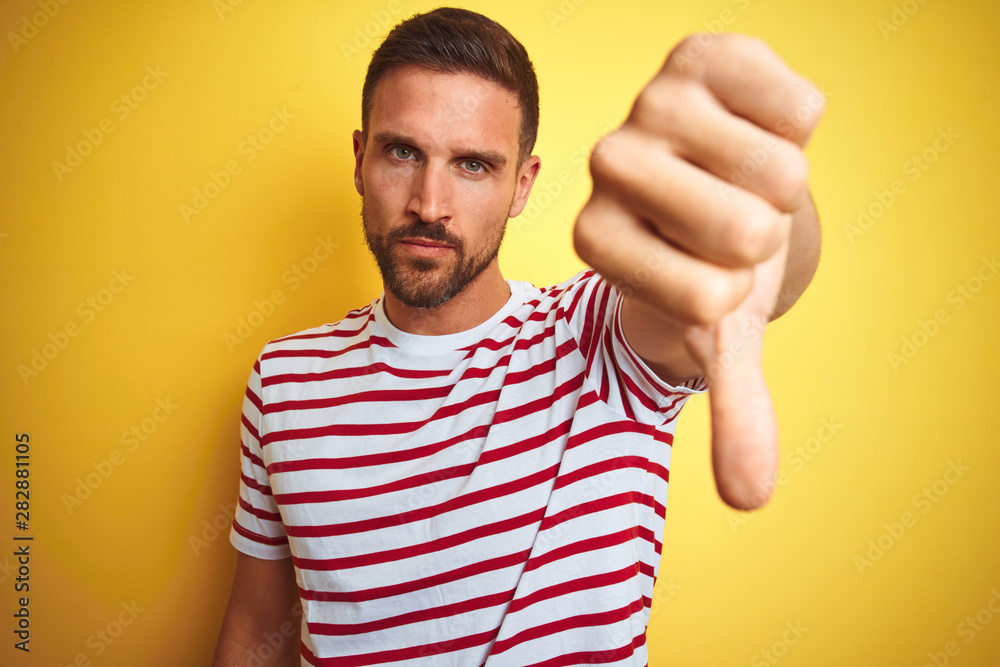Young handsome man wearing casual red striped t-shirt over yellow isolated background looking unhappy and angry showing rejection and negative with thumbs down gesture. Bad expression. Young handsome man wearing casual red striped t-shirt over yellow isolated background looking unhappy and angry showing rejection and negative with thumbs down gesture. Bad expression.