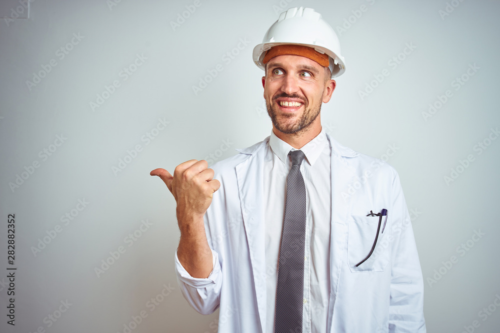 Young handsome engineer man wearing safety helmet over isolated background smiling with happy face looking and pointing to the side with thumb up. Young handsome engineer man wearing safety helmet over isolated background smiling with happy face looking and pointing to the side with thumb up.