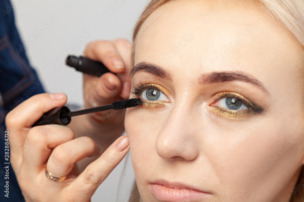 Makeup artist puts on an oriental-style make-up with gold and green shades of a young attractive blonde girl, paints her eyelashes with mascara, hold on the upper eyelid, holding a brush. Makeup artist puts on an oriental-style make-up with gold and green shades of a young attractive blonde girl, paints her eyelashes with mascara, hold on the upper eyelid, holding a brush.
