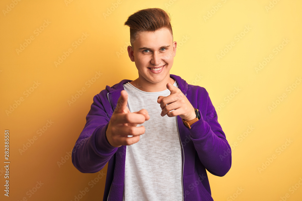 Young handsome man wearing purple sweatshirt standing over isolated yellow background pointing fingers to camera with happy and funny face. Good energy and vibes. Young handsome man wearing purple sweatshirt standing over isolated yellow background pointing fingers to camera with happy and funny face. Good energy and vibes.