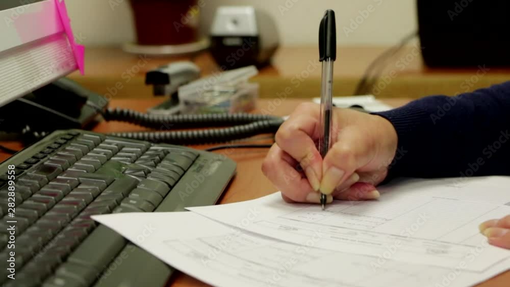 Detalle de una mano de mujer escribiendo con un bolígrafo en documentos de oficina frente a una computadora