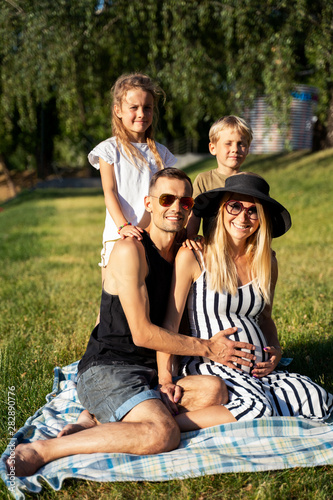 Picture of father and pregnant mother with children sitting on plaid on picnic on summer day © snedorez