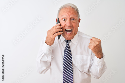 Senior grey-haired businessman talking on the smartphone over isolated white background screaming proud and celebrating victory and success very excited, cheering emotion © Krakenimages.com