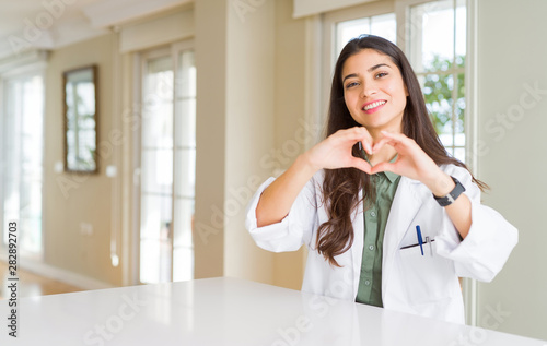 Young woman wearing medical coat at the clinic as therapist or doctor smiling in love showing heart symbol and shape with hands. Romantic concept.