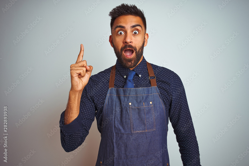 Young indian employee man wearing apron uniform standing over isolated white background pointing finger up with successful idea. Exited and happy. Number one. Young indian employee man wearing apron uniform standing over isolated white background pointing finger up with successful idea. Exited and happy. Number one.