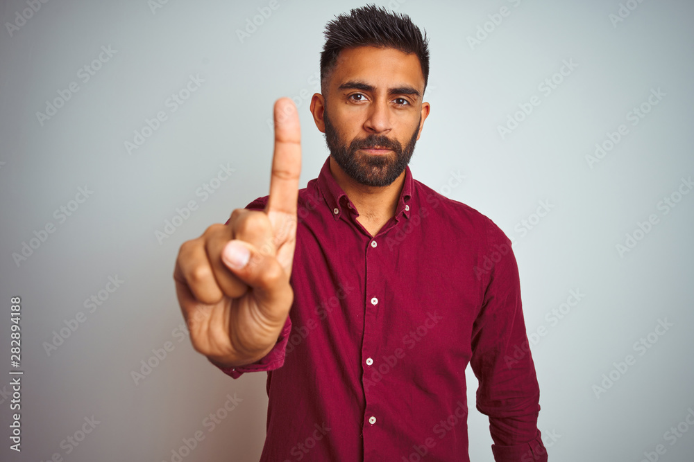 Young indian man wearing red elegant shirt standing over isolated grey background Pointing with finger up and angry expression, showing no gesture Young indian man wearing red elegant shirt standing over isolated grey background Pointing with finger up and angry expression, showing no gesture