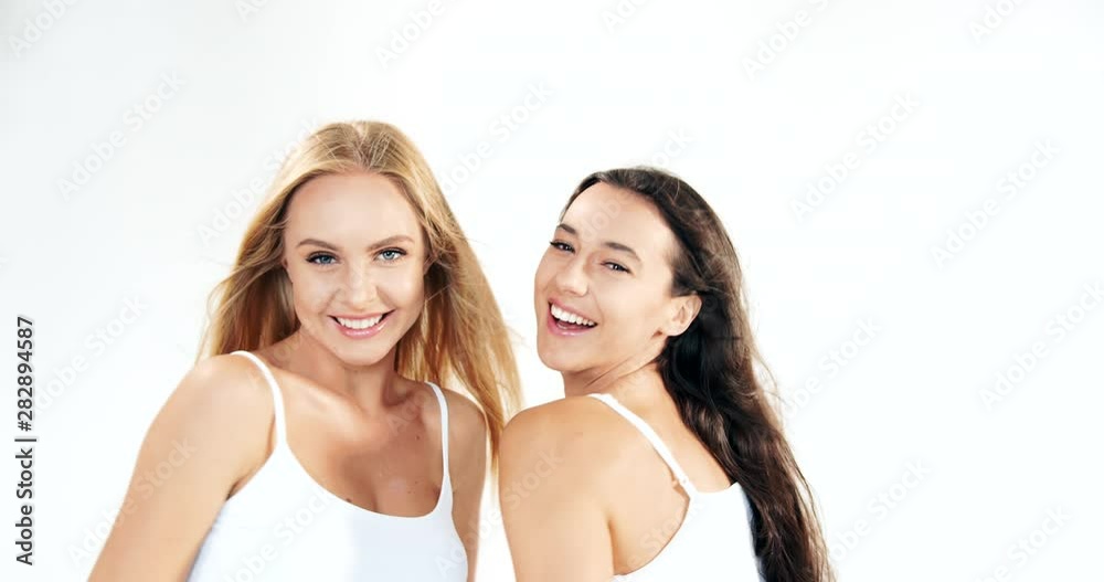 Two attractive dark-haired and blonde ladies in white tops dancing on white background, smiling and having fun, beauty Two attractive dark-haired and blonde ladies in white tops dancing on white background, smiling and having fun, beauty